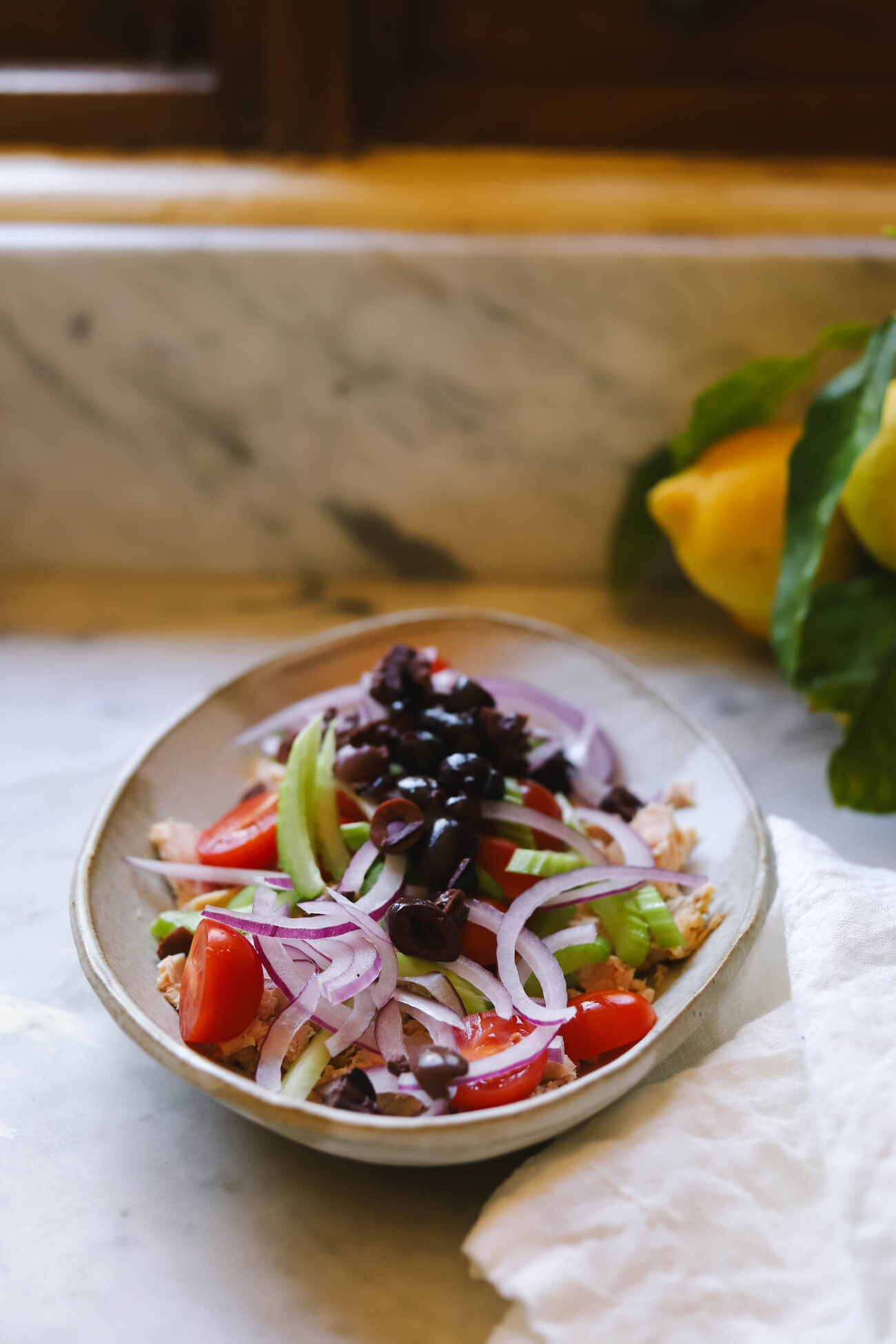 A stoneware bowl filled with tuna, cherry tomatoes, celery, red onion, and kalamata olives.
