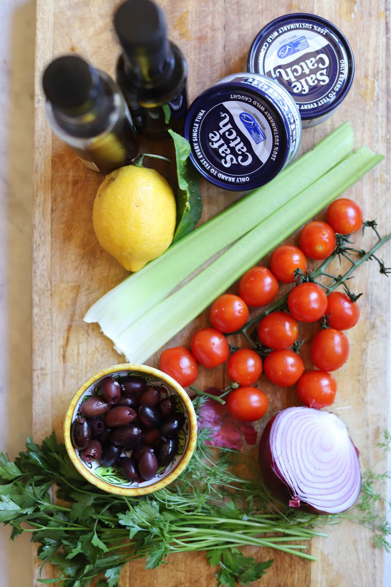 Mediterranean Tuna Salad Ingredients ngredients for Mediterranean tuna salad including albacore tuna, cherry tomatoes, celery, red onion, kalamata olives, parsley, lemon, olive oil, and red wine vinegar on a wooden board.